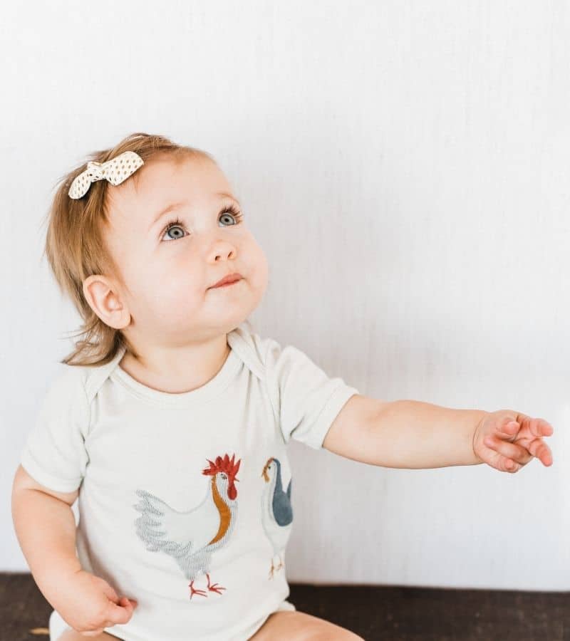 Baby Girl with a Bow Wearing the Organic Applique One Piece with the Chicken Applique by Milkbarn Kids