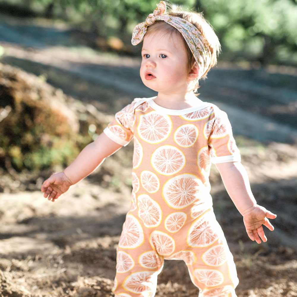 Little Baby Girl in a Field Wearing the Organic Cotton Romper in the Grapefruit Print and the Bamboo Headband in the Rose Floral Print by Milkbarn Kids