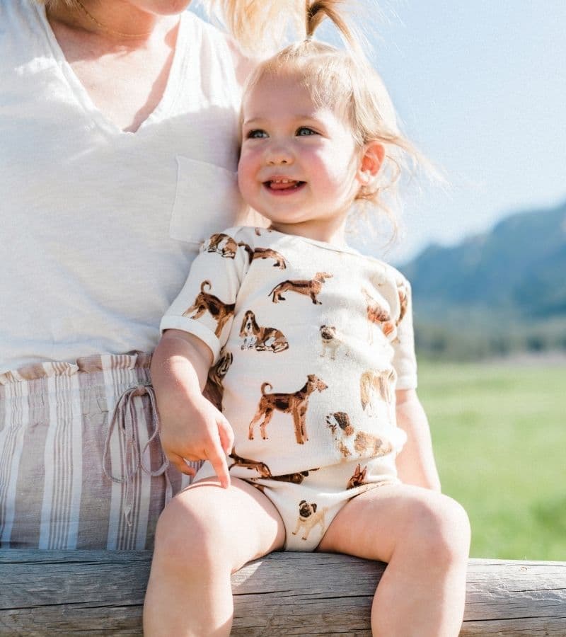Baby Girl with her Mom by a green field wearing the Organic Cotton One Piece in the Natural Dog Print