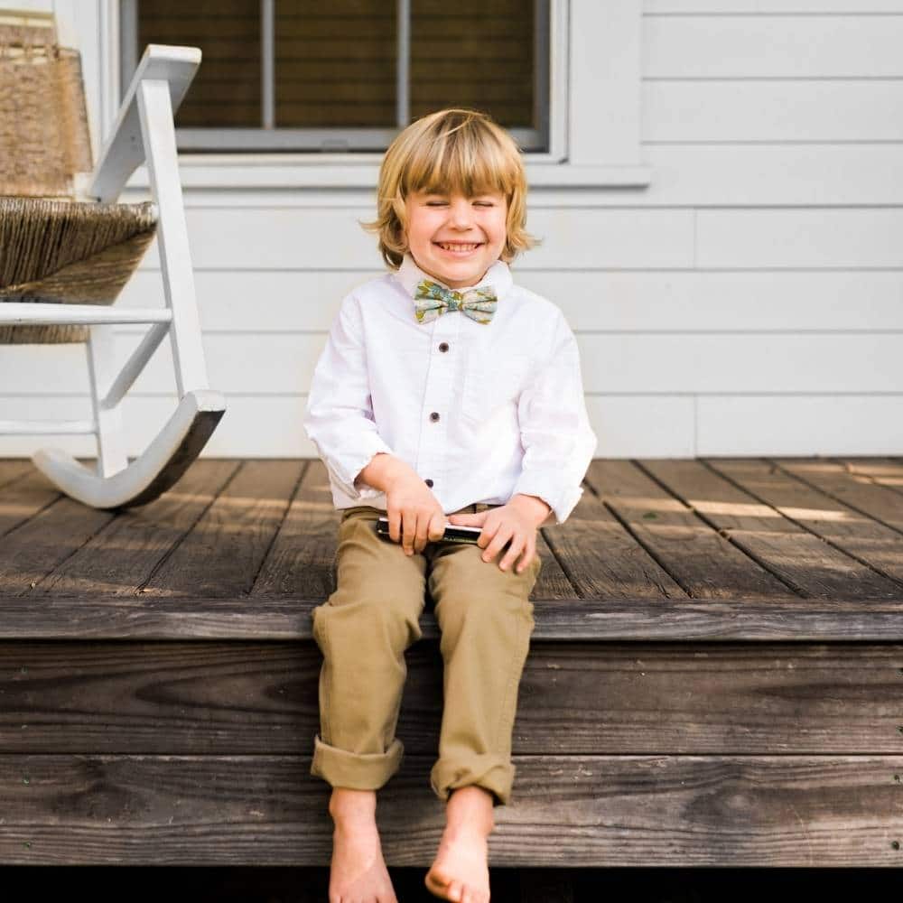 Boy Sitting on a Porch Laughing Wearing Blue Floral Bow Tie by Milkbarn Kids