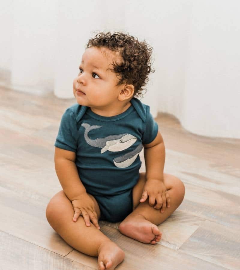 Baby Boy Sitting on a Wood Floor Wearing the Organic Cotton Applique Whale One Piece by Milkbarn Kids