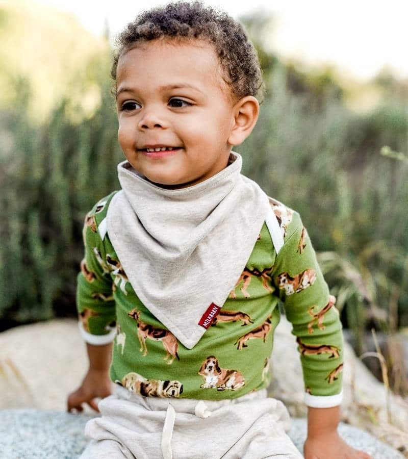 Baby Boy Sitting on a Rock in a Field Wearing the Organic Cotton Kerchief Bib by Milkbarn Kids