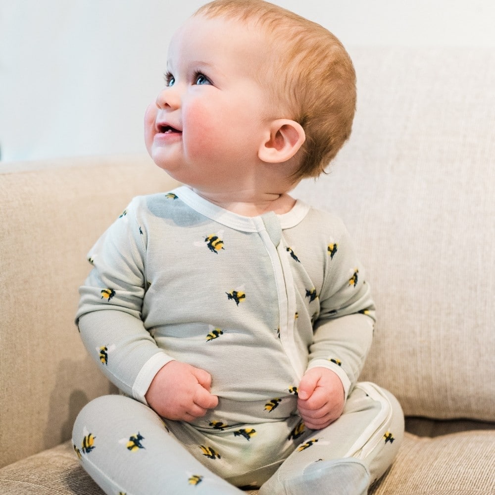 Baby sitting on a beige couch looking off smiling wearing the Bumblbee Bamboo Zipper Footed Romper by Milkbarn Kids