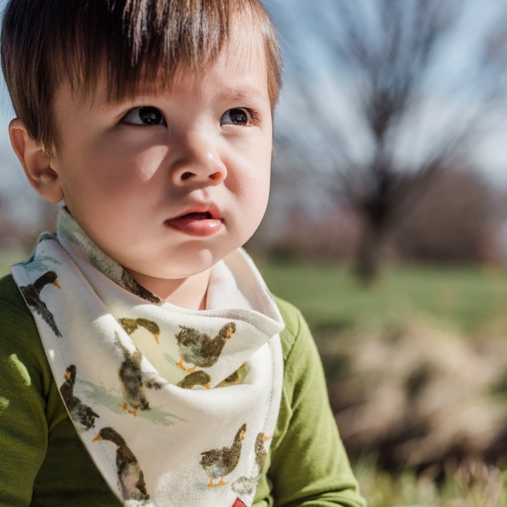 Baby boy outside in a green shirt wearing the Duck Organic Cotton Kerchief Bib by Milkbarn Kids