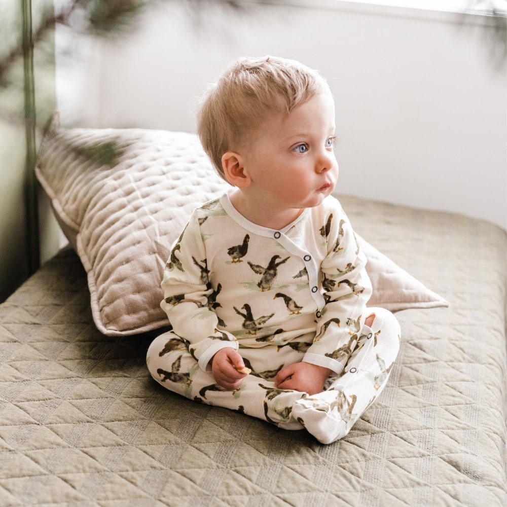 Baby boy sitting on a bed wearing the Duck Organic Cotton Snap Footed Romper by Milkbarn Kids