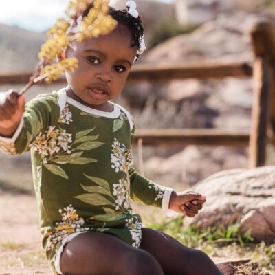 Baby Girl in a field holding yellow flowers wearing the Green Floral Bamboo Long Sleeve One Piece by Milkbarn Kids