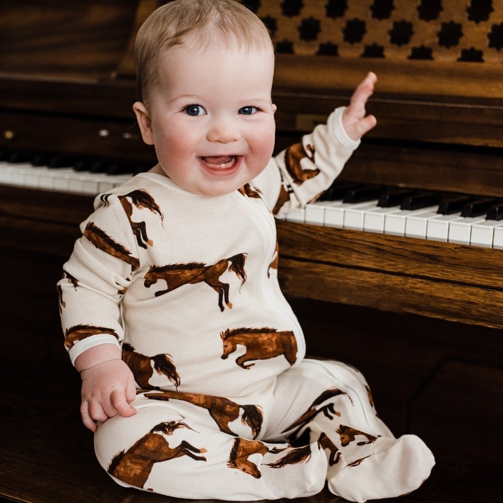 Baby sitting at a piano smiling wearing the Natural Horse Organic Cotton Zipper Footed Romper by Milkbarn Kids