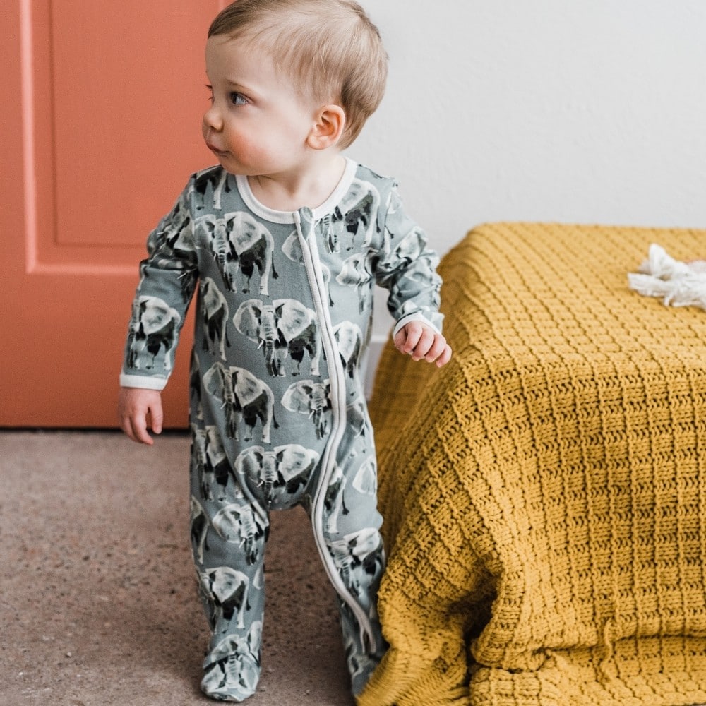 Baby boy at the foot of a bed wearing the Organic Cotton Zipper Footed Romper in the Grey Elephant print by Milkbarn Kids