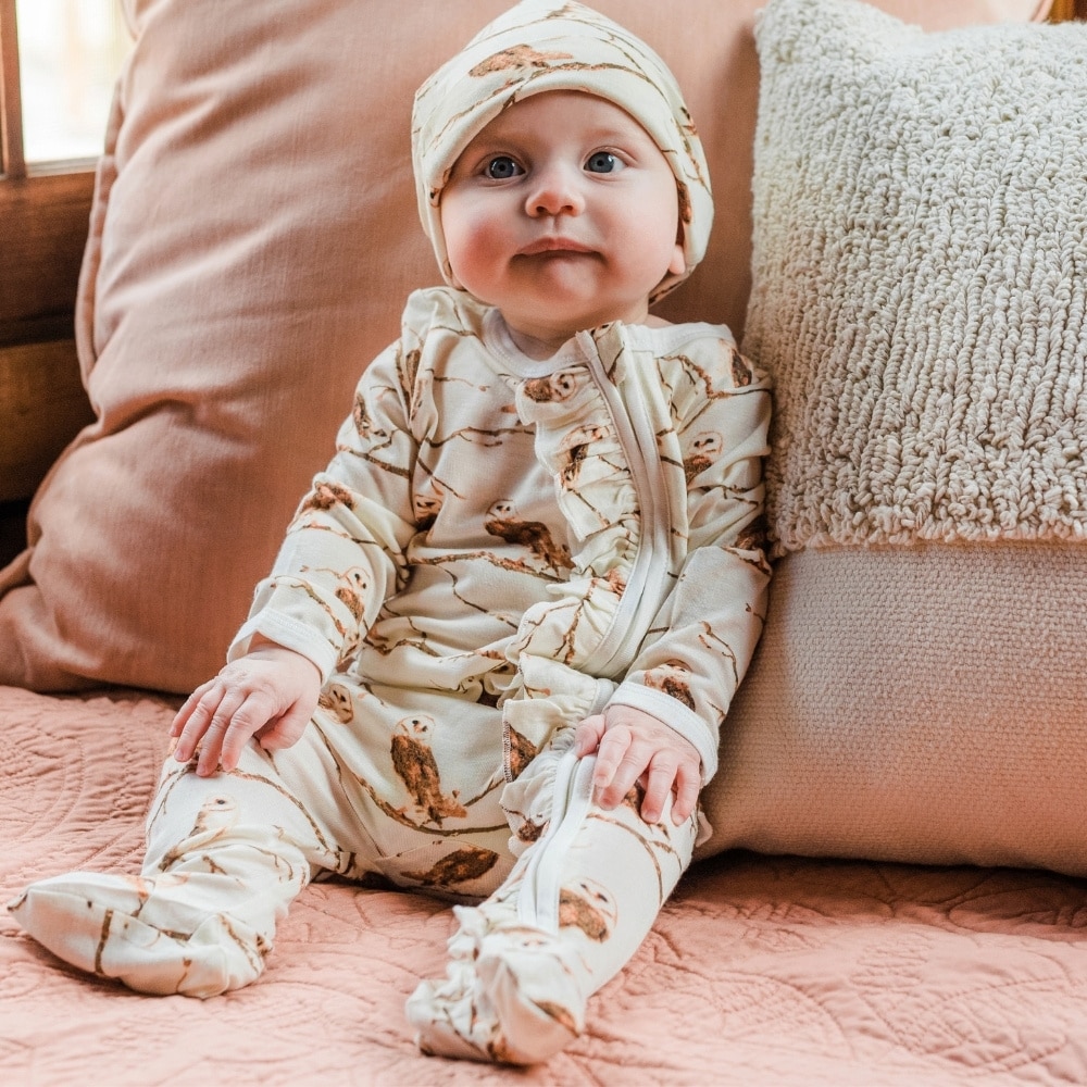 Baby propped up on a pink velvet couch and pillow wearing the owl Bamboo Ruffle Zipper Footed Romper and matching Owl Bamboo Knotted Hat by Milkbarn Kids