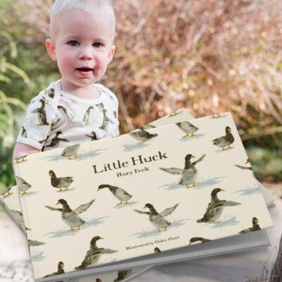 Little Boy outside in a garden wearing the duck print short sleeve one piece by Milkbarn Kids with the matching book Little Huck by Rory Feek