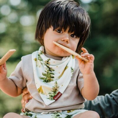 Baby boy sitting up eating a graham cracker while wearing the Camping Bamboo Kerchief Bib by Milkbarn