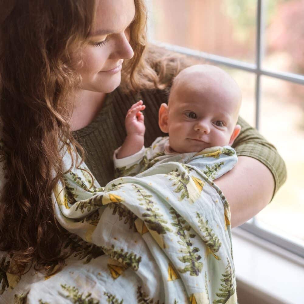 Mom cradles her son while standing near a window. Her son is wrapped in the unfolded Camping Bamboo Muslin Swaddle Blanket by Milkbarn
