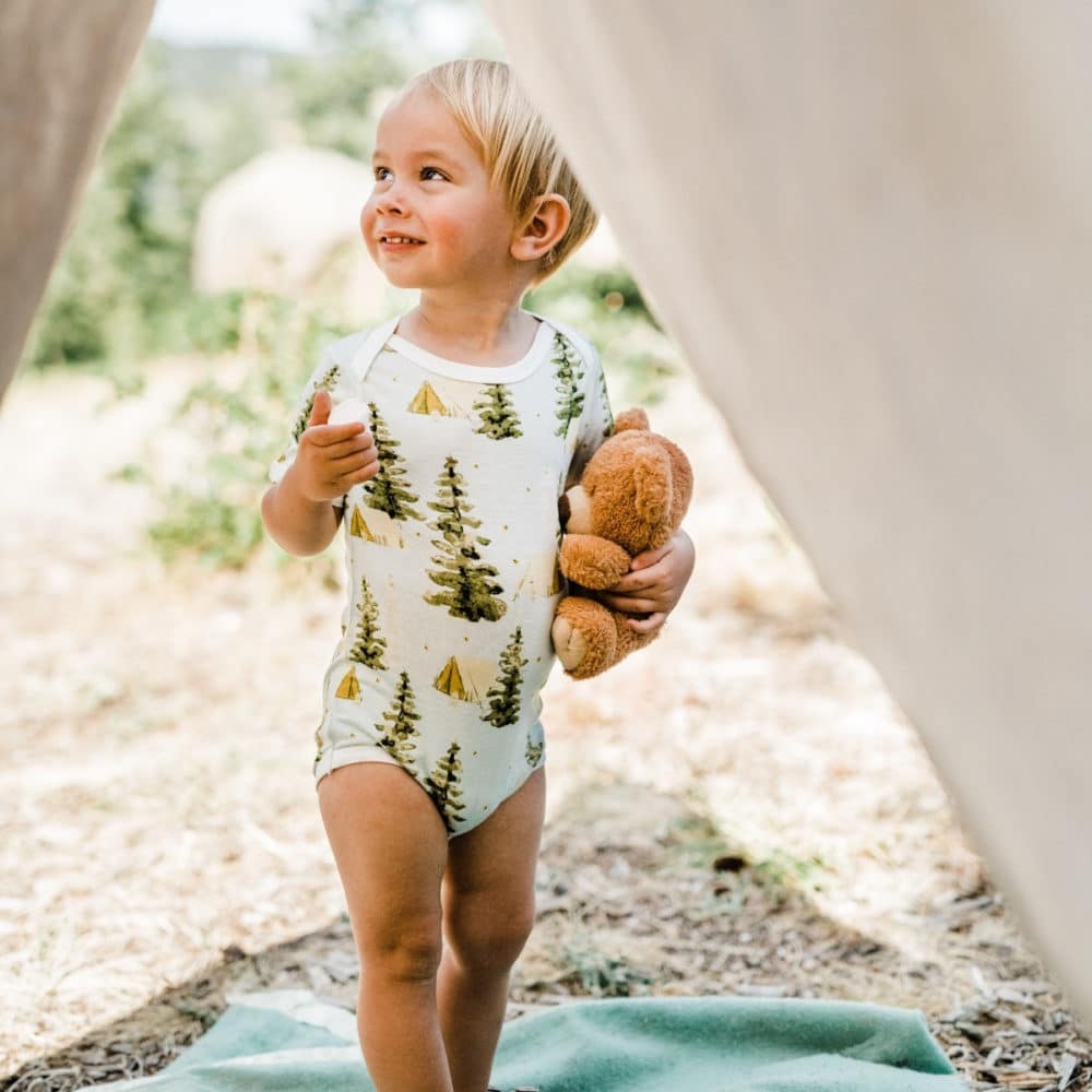 Baby boy standing in a tent entrance holding a teddy bear wearing the Camping Bamboo Short Sleeve One Piece by Milkbarn Kids