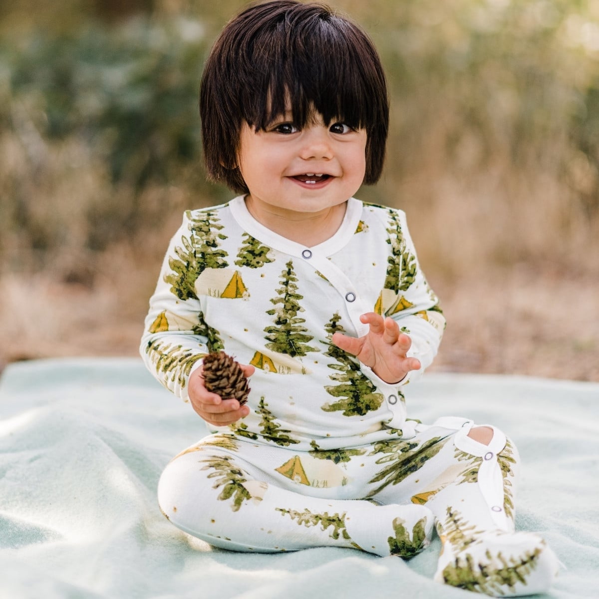 Baby sitting on a blanket holding a pinecone and wearing the Camping Bamboo Snap Footed Romper by Milkbarn Kids