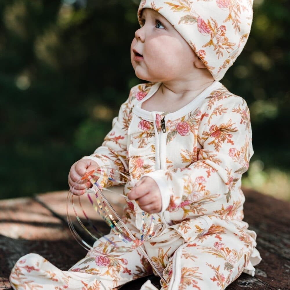 Baby Girl Sitting Outside Wearing the Ruffle Zipper Footed Romper and Matching Knotted Beanie Hat in the Vintage Floral Print by Milkbarn Kids