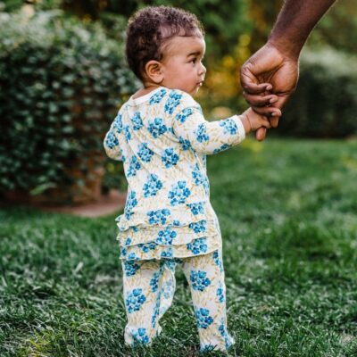 Baby girl on green grass holding her Dad's hand wearing the Sky Floral Bamboo Ruffle Zipper Footed Romper by Milkbarn Kids