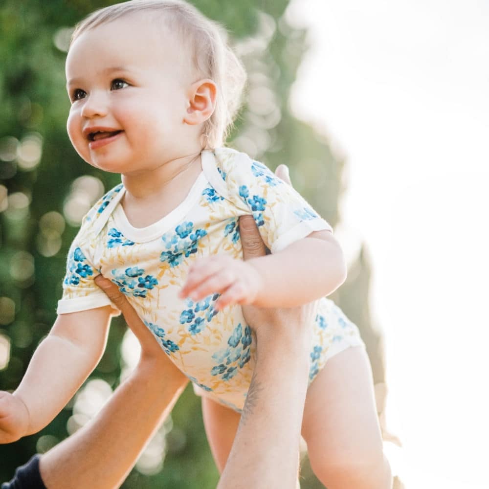 Baby Girl being Held up in the Sky wearing the Sky Floral Bamboo Short Sleeve One Piece by Milkbarn Kids