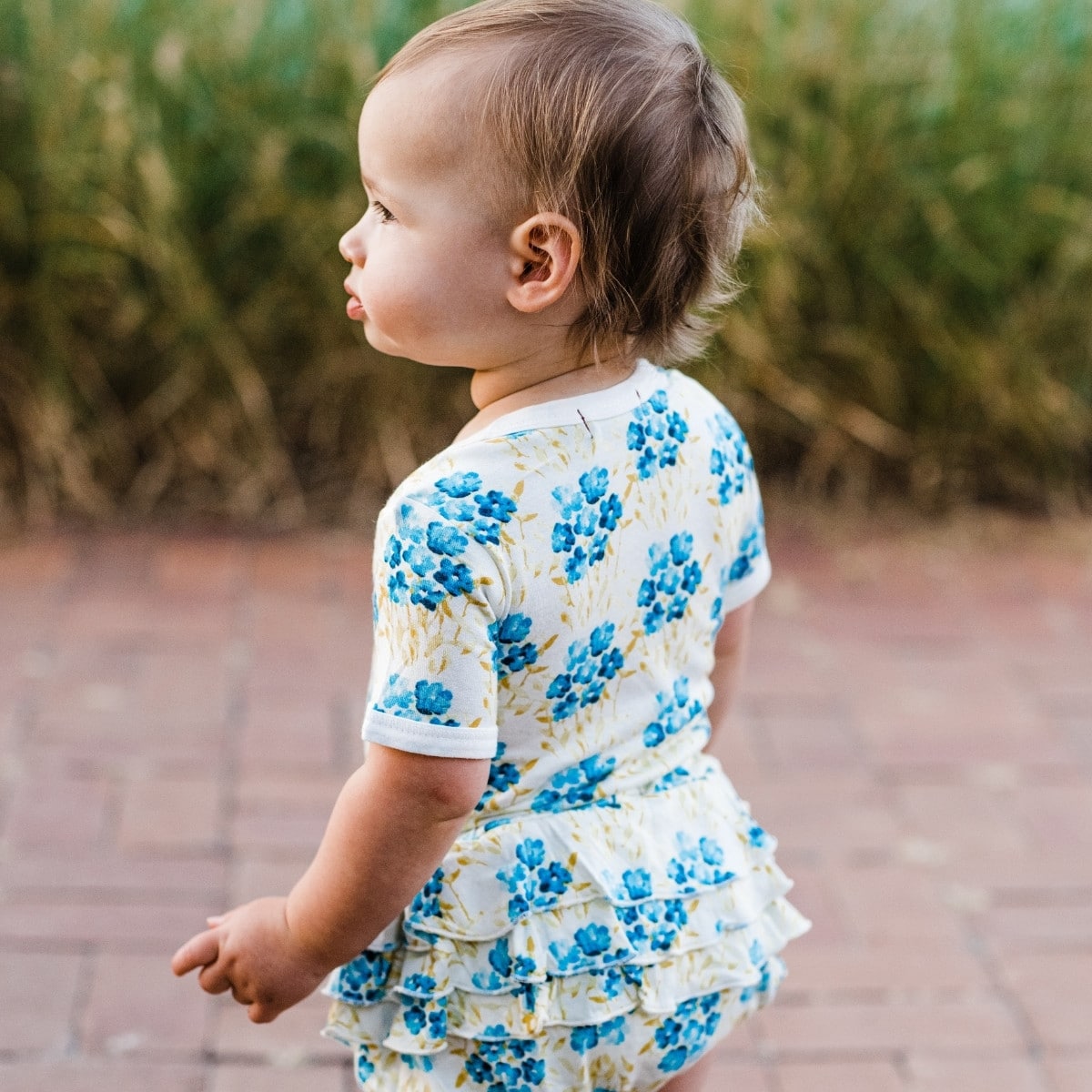 Baby girl outsideon a red brick path turned away wearing the Sky Floral Bamboo Short Sleeve One Piece and matching ruffle bloomers by Milkbarn Kids