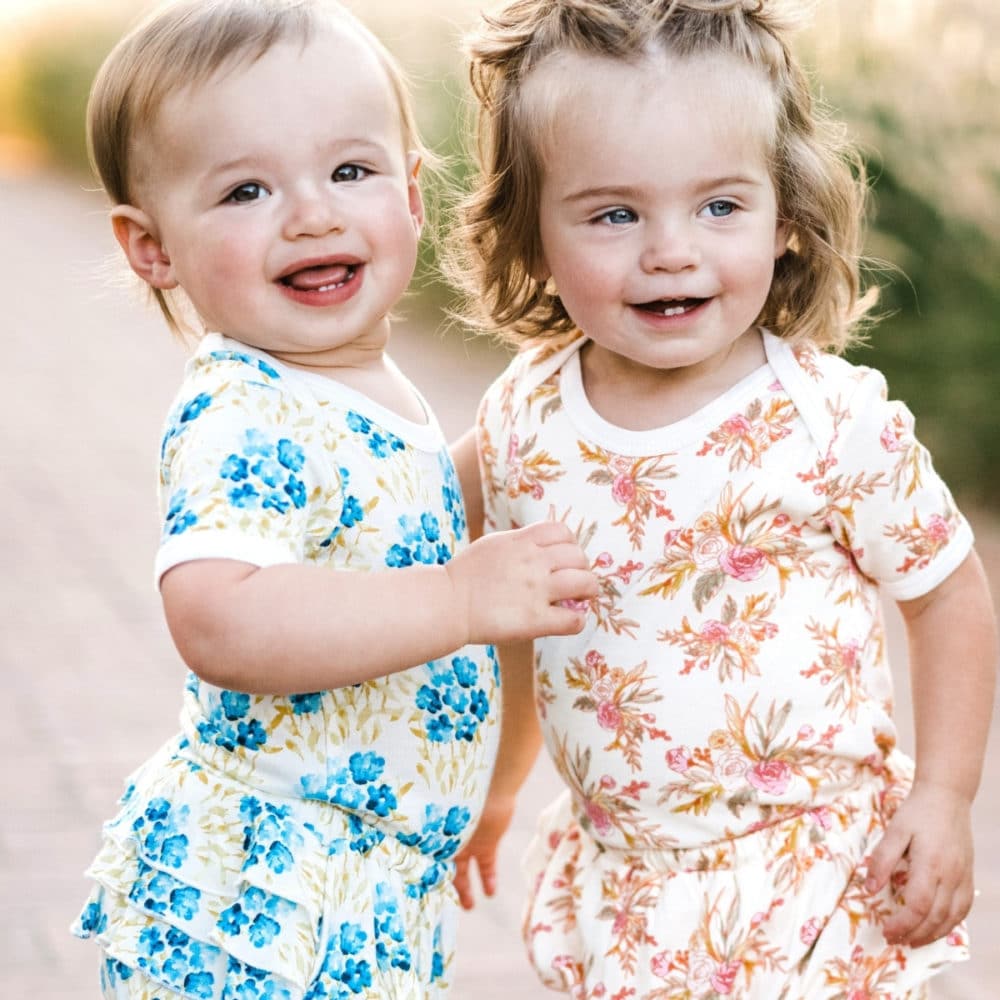 Two baby girls together smiling outside wearing matching outfits in Sky Floral and Vintage Floral Short Sleeve One Pieces and Ruffle Bloomers by Milkbarn Kids