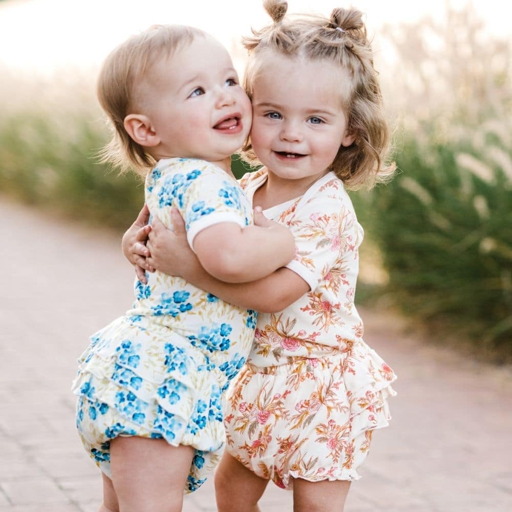 Two Baby Girls on a Brick Path Hugging and Wearing the Sky and Vintage Floral Ruffle Bloomers by Milkbarn Kids