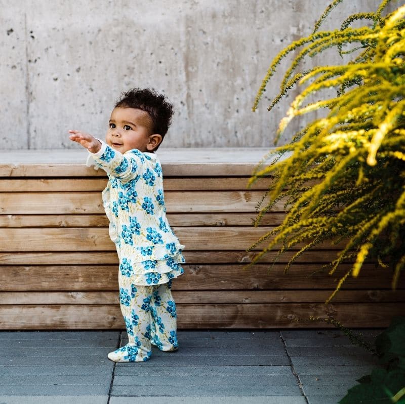 Baby girl reaching out wearing the Blue Sky floral Bamboo Ruffle Footed Romper by Milkbarn Kids