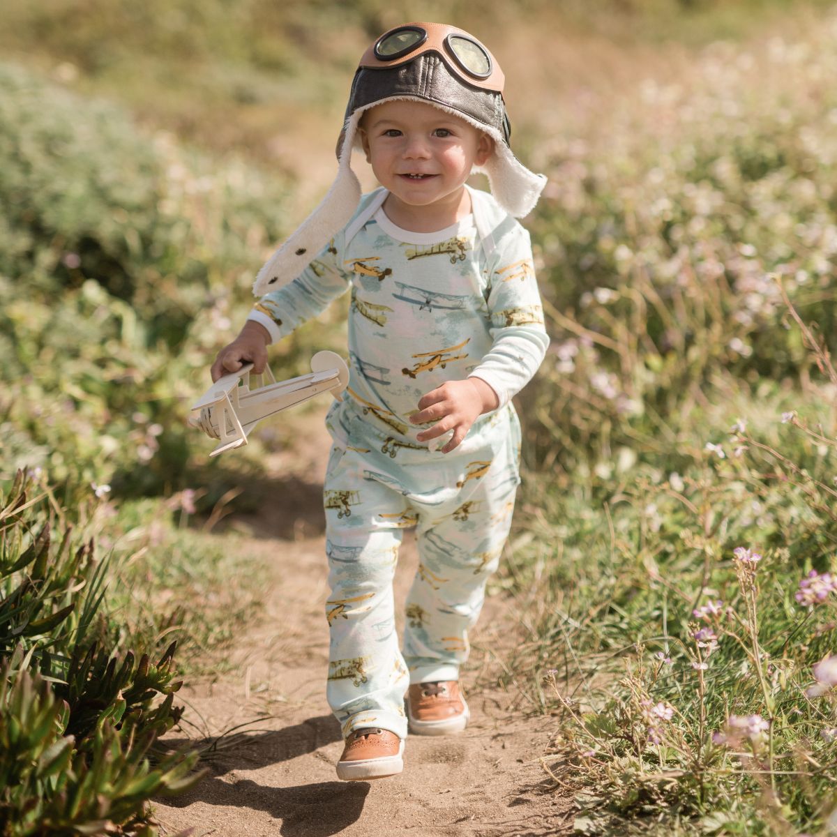 Baby boy in a field by the coast running with a toy vintage airplane while wearing the Vintage Plane Organic Jogger Pant by Milkbarn