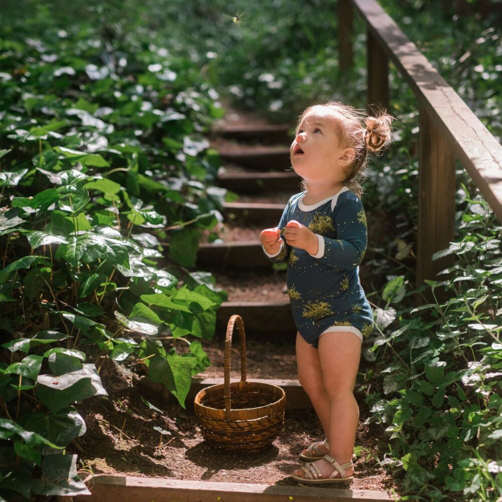 Baby girls standing on forest steps looking up at a firefly while wearing the long sleeve bamboo one piece in the firefly print by Milkbarn