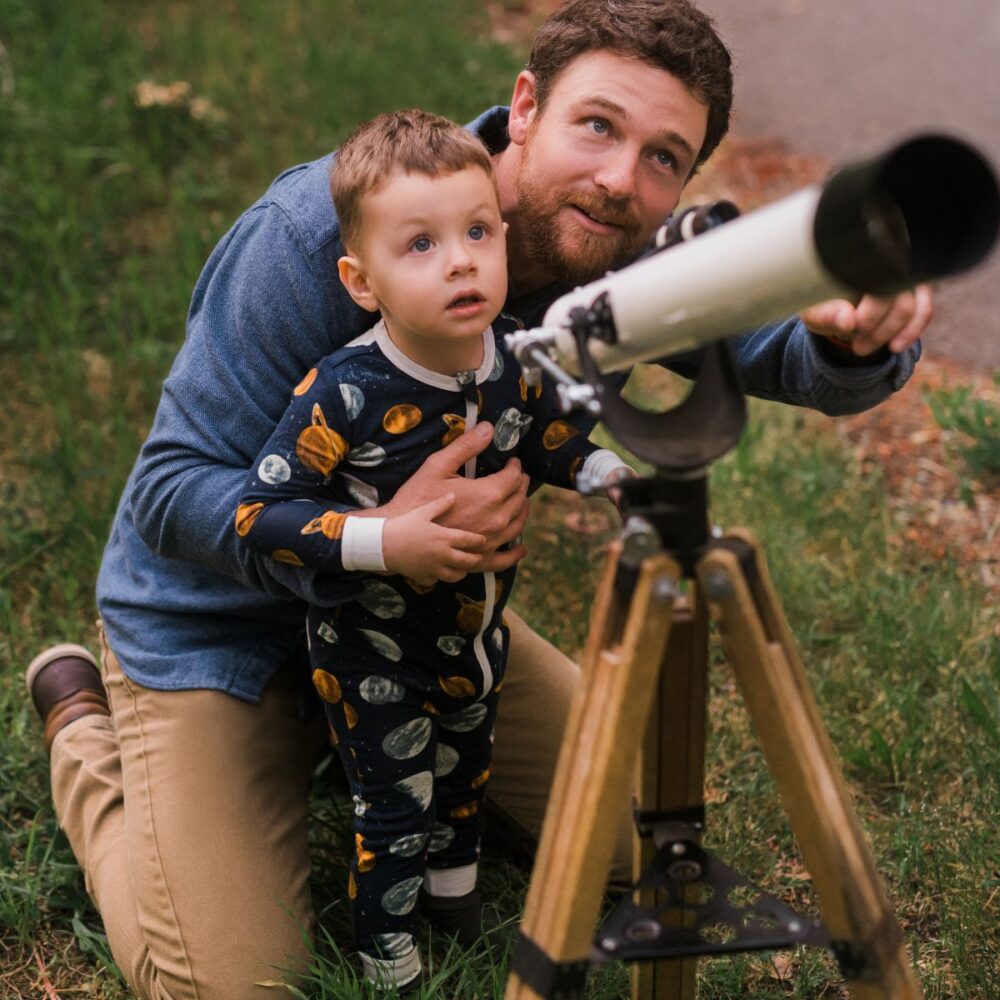 Dad kneeling down and hugging his baby boy while helping him look through a telescope trying to find the planets, stars and space while the son wears the Planets Bamboo Zipper Pajama by Milkbarn