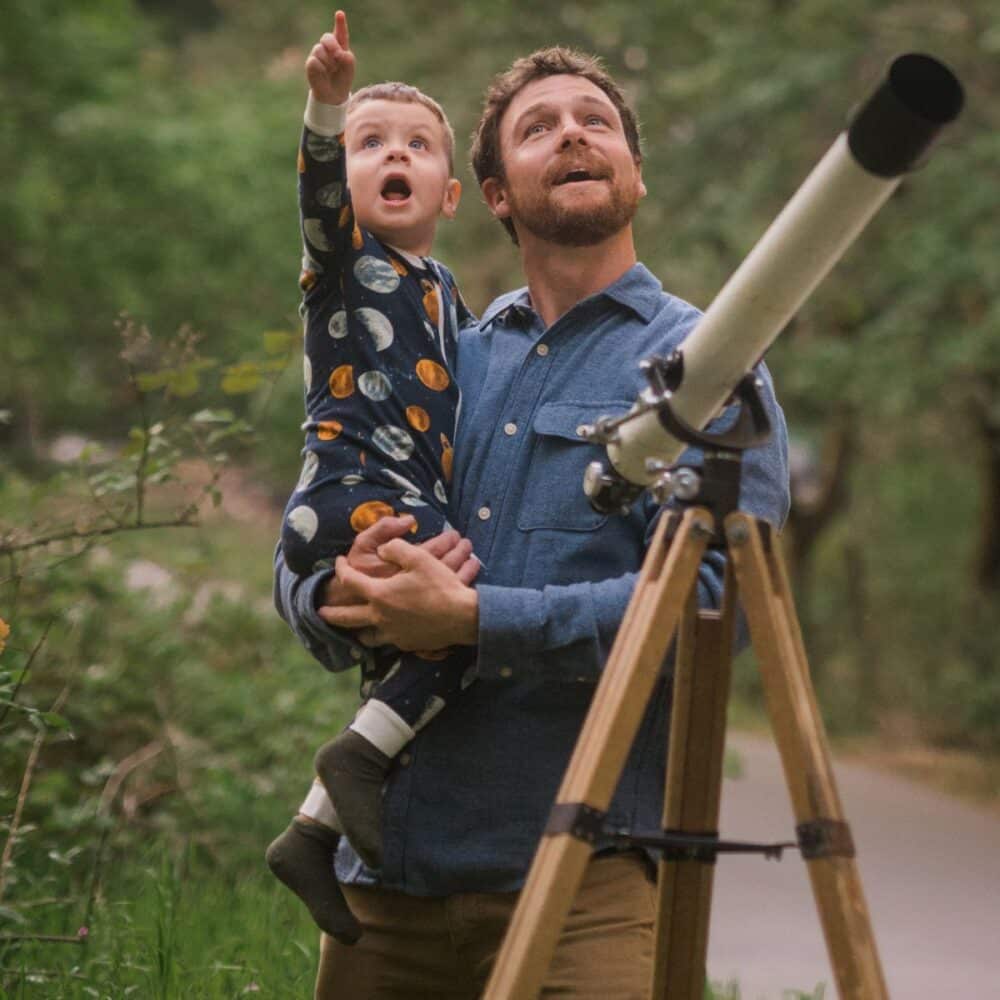 Baby boy and Dad outside with a telescope looking up at the sky trying to find the planets and space while wearing the Planets Bamboo Zipper Pajama by Milkbarn