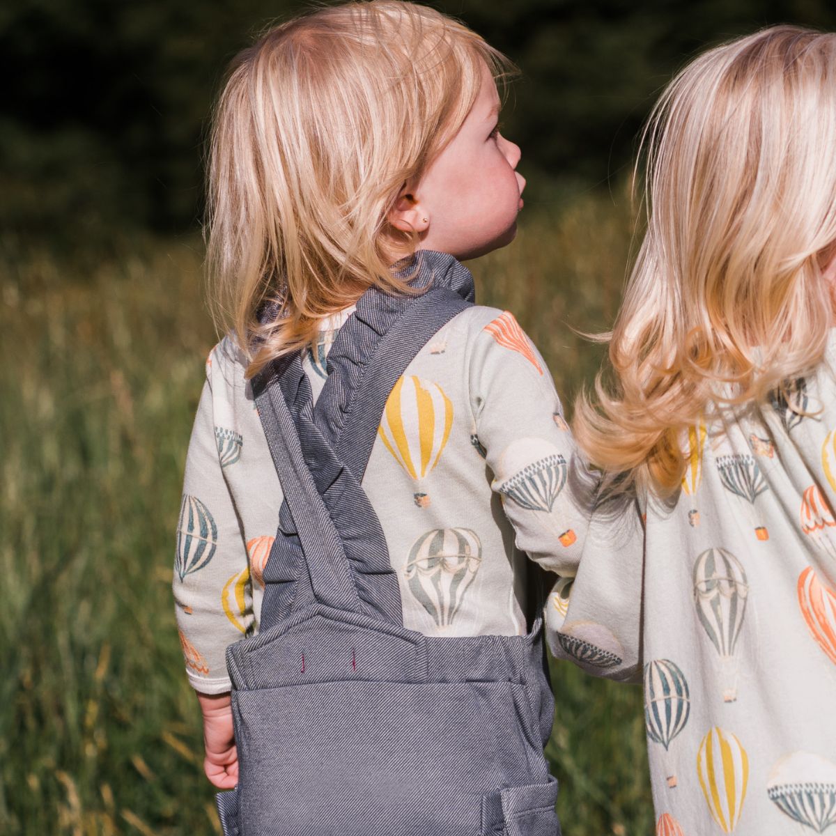 Baby girl holding hands with her sister and looking up into the sky while wearing the Vintage Balloons Organic Long Sleeve One Piece and Denim Ruffle Overalls by Milkbarn