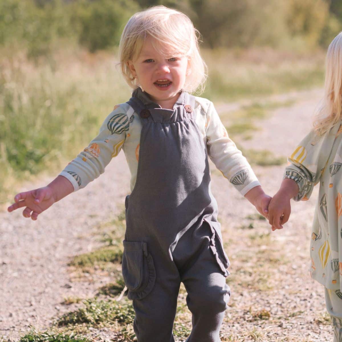 Baby girl holding hands with her sister walking along a path wearing the Vintage Balloons Organic Long Sleeve One Piece and Denim Ruffle Overalls by Milkbarn