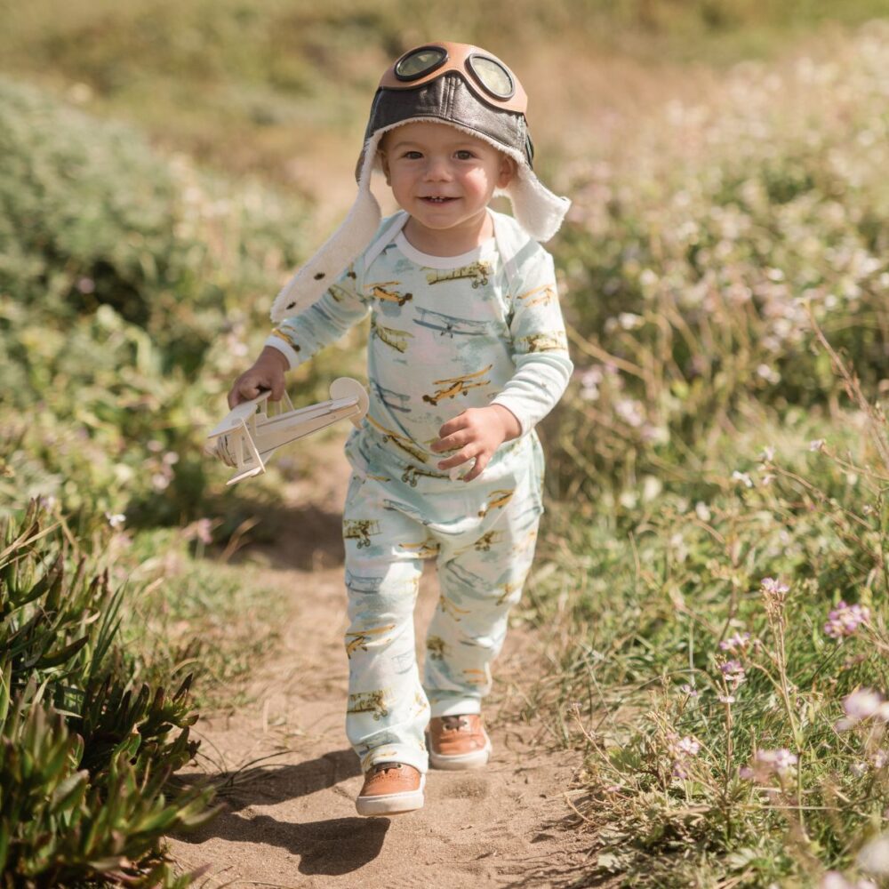 Baby boy running in a field near the ocean wearing an aviator hat and holding a toy vintage airplane while wearing the Vintage Planes Organic Long Sleeve One Piece and Jogger pants by Milkbarn