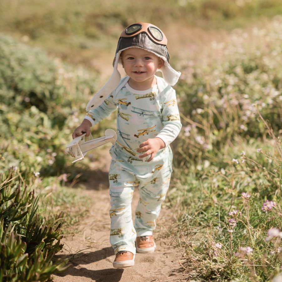 Baby boy wearing an aviator hat and running off the coast of northern California wearing an outfit with the Vintage Planes organic cotton print by Milkbarn and holding a wooden biplane.