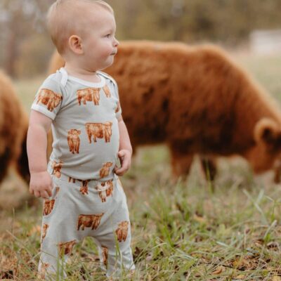 Baby boy in a cow field standing next to brown cows wearing the bamboo short sleeve one piece and jogger pants in the matching Highland Cow print by Milkbarn