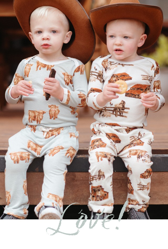 Two baby boys sit together wearing cowboy hats and each wearing the long sleeve one piece and jogger pants in the Highland Cow print and the Homestead print