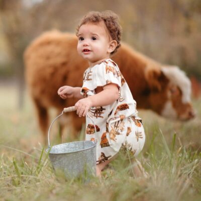 Baby girl in an open field with highland cows carrying a bucket wearing a dress and bloomer set by Milkbarn in the Homestead organic cotton print.