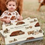 Baby girl in a cow field wearing the Homestead organic cotton print dress and bloomers that match the book cover of Mr. Harvey's Barn, the children's book by Milkbarn Kids and illustrated by Haley Hunt