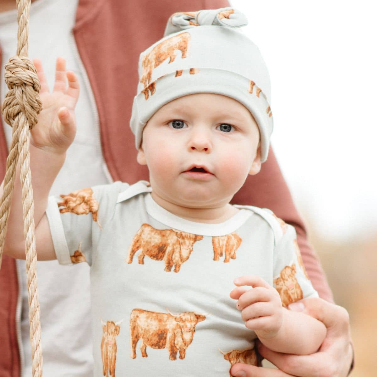 Baby boy on a swing being held by his Dad wearing the bamboo knotted beanie hat and short sleeve one piece in the highland cow print by Milkbarn Kids