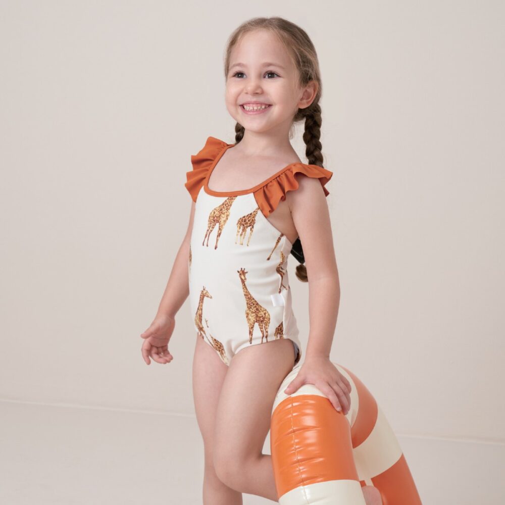 Toddler girl wearing the Giraffe Ruffle Cross Back One Piece Swimsuit Lifestyle while holding a round orange pool floatie