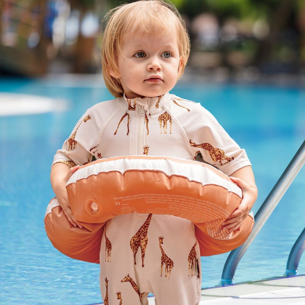 Toddler boy by the swimming pool with a round floatie wearing the Giraffe Zipper Short Sleeve Shortall Swimsuit.