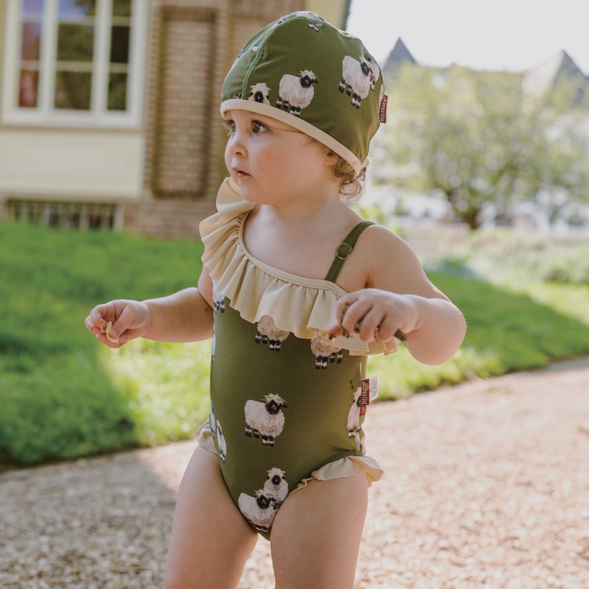 Baby girl by the pool and green grass wearing the Valais Sheep Ruffle Off Shoulder One Piece Swimsuit by Milkbarn