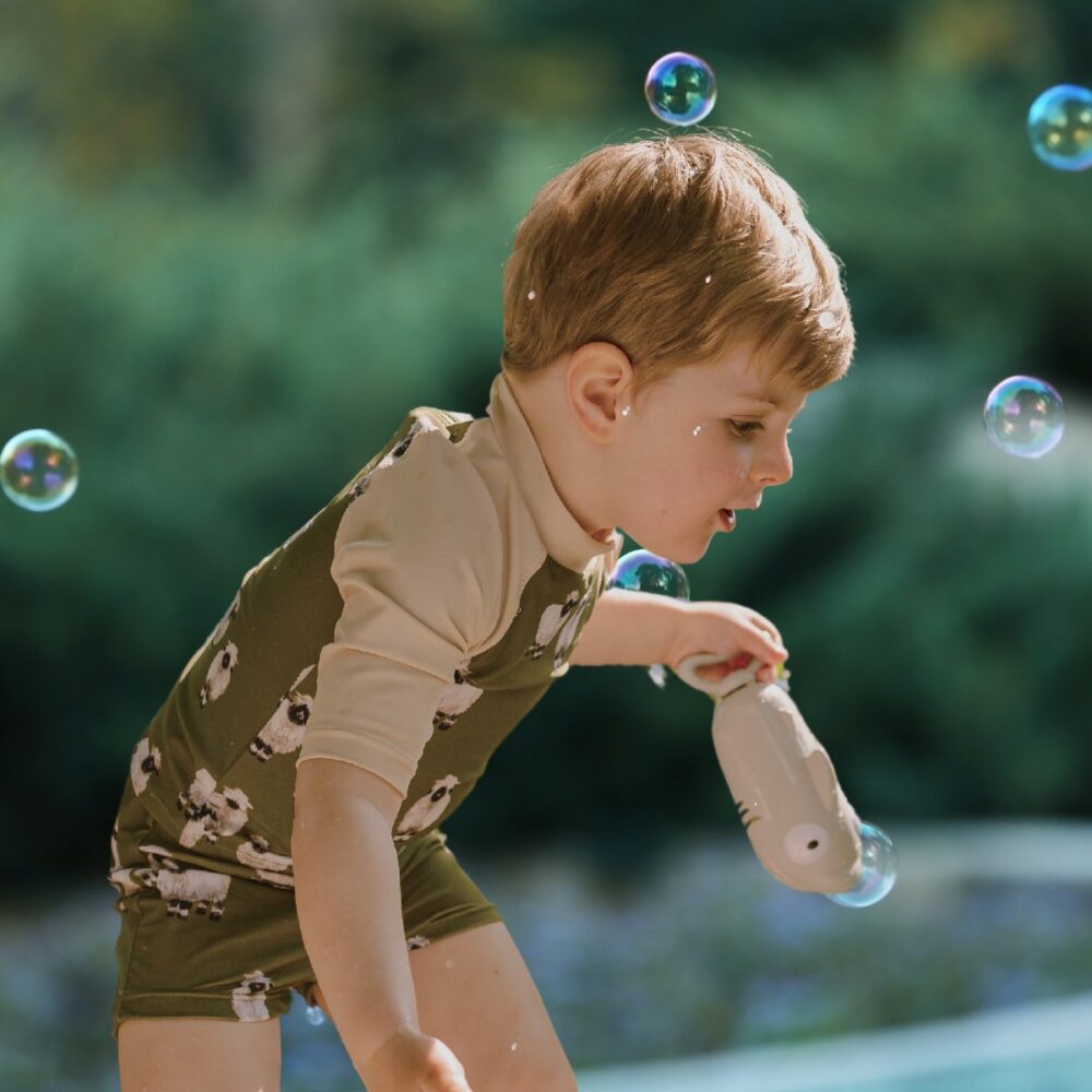 Toddler boy playing by the pool and blowing bubbles while wearing the Valais Sheep Zipper Top and Swim Trunks Lifestyle 4