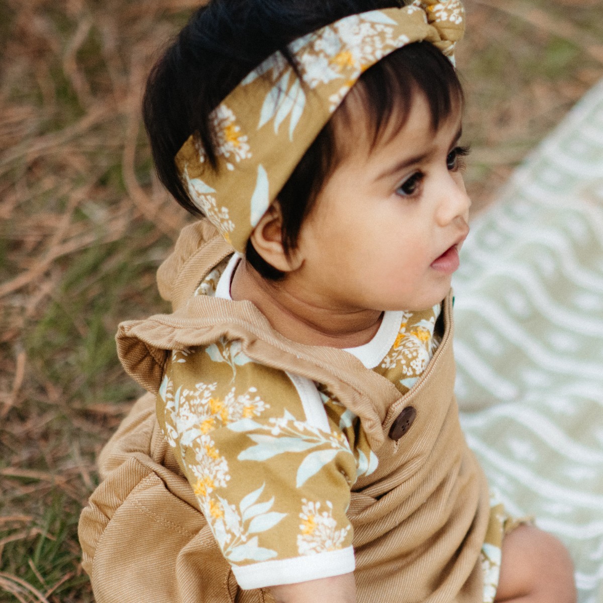Baby girl sitting on a picnic blanket wearing the Gold Floral organic cotton Knotted Headband by Milkbarn