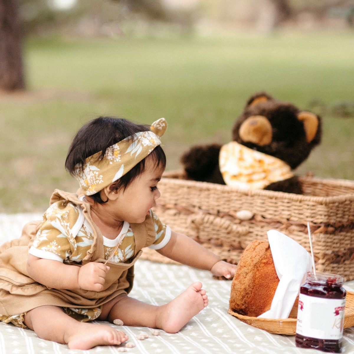 Baby girl sitting on a picnic blanket with a picnic basket with a stuffed bear and wearing the Gold Floral organic cotton Knotted Headband by Milkbarn