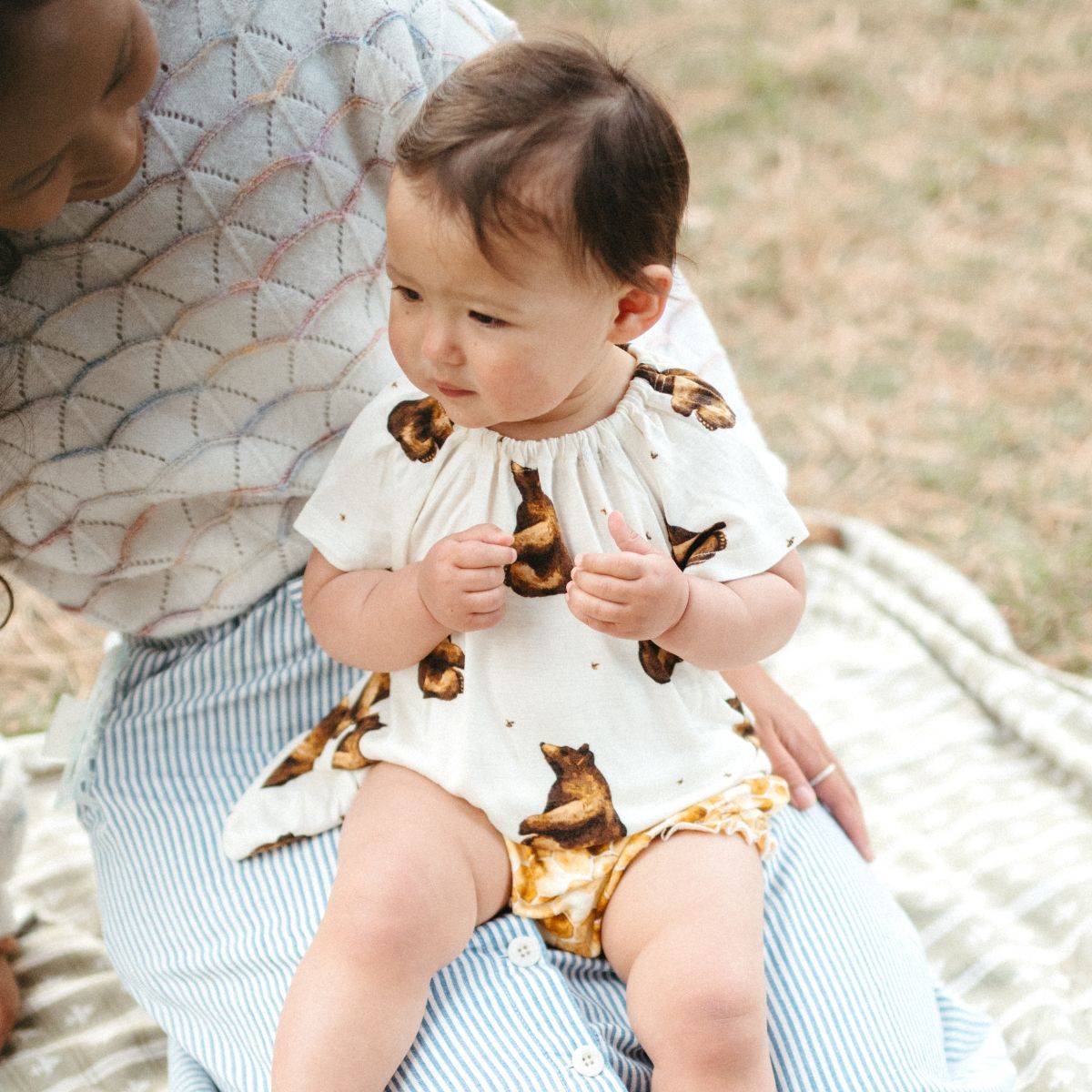 Baby girl being held on her mother's lap at a park while wearing the bamboo Honey Bear Dress top and Honeycomb bamboo Bloomers by Milkbarn