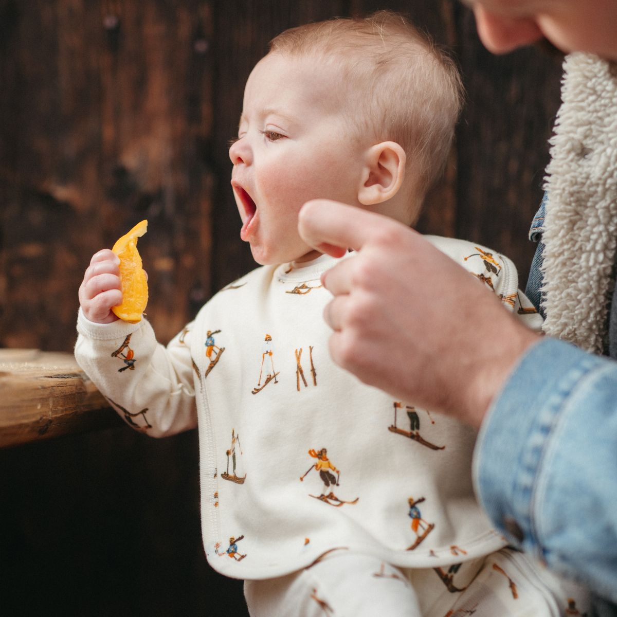 Dad in a jean jacket holding a baby outside trying to eat an orange slice while wearing the Vintage Natural Ski Organic Cotton Traditional Bib by Milkbarn Kids