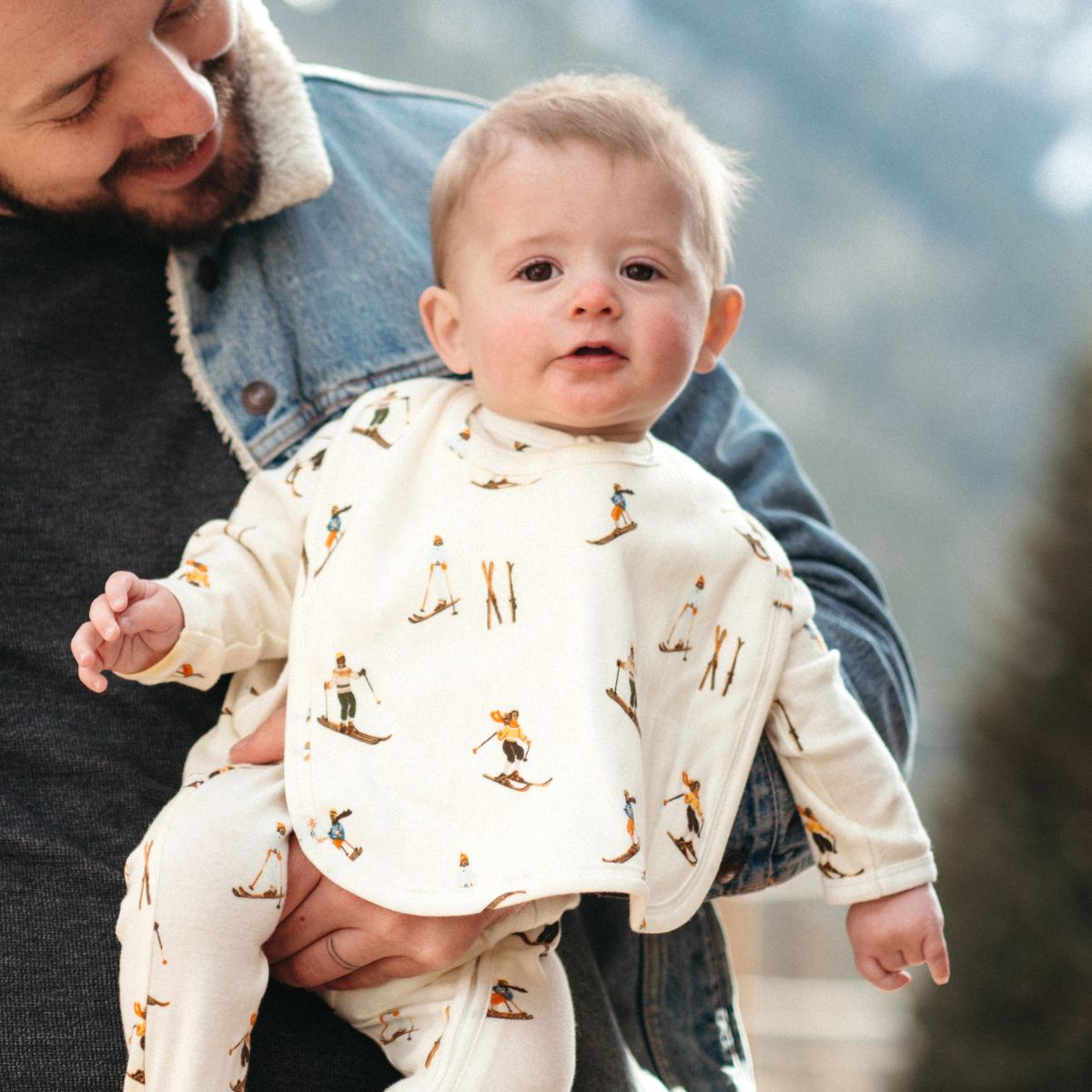 Dad in a jean jacket holding a baby outside wearing the Vintage Natural Ski Organic Cotton Traditional Bib by Milkbarn Kids
