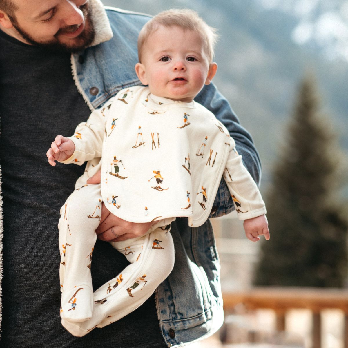 Dad and son in the snow with the baby wearing the Vintage Natural Ski print on the organic cotton Zipper Footed Romper Lifestyle and matching traditional bib by Milkbarn Kids