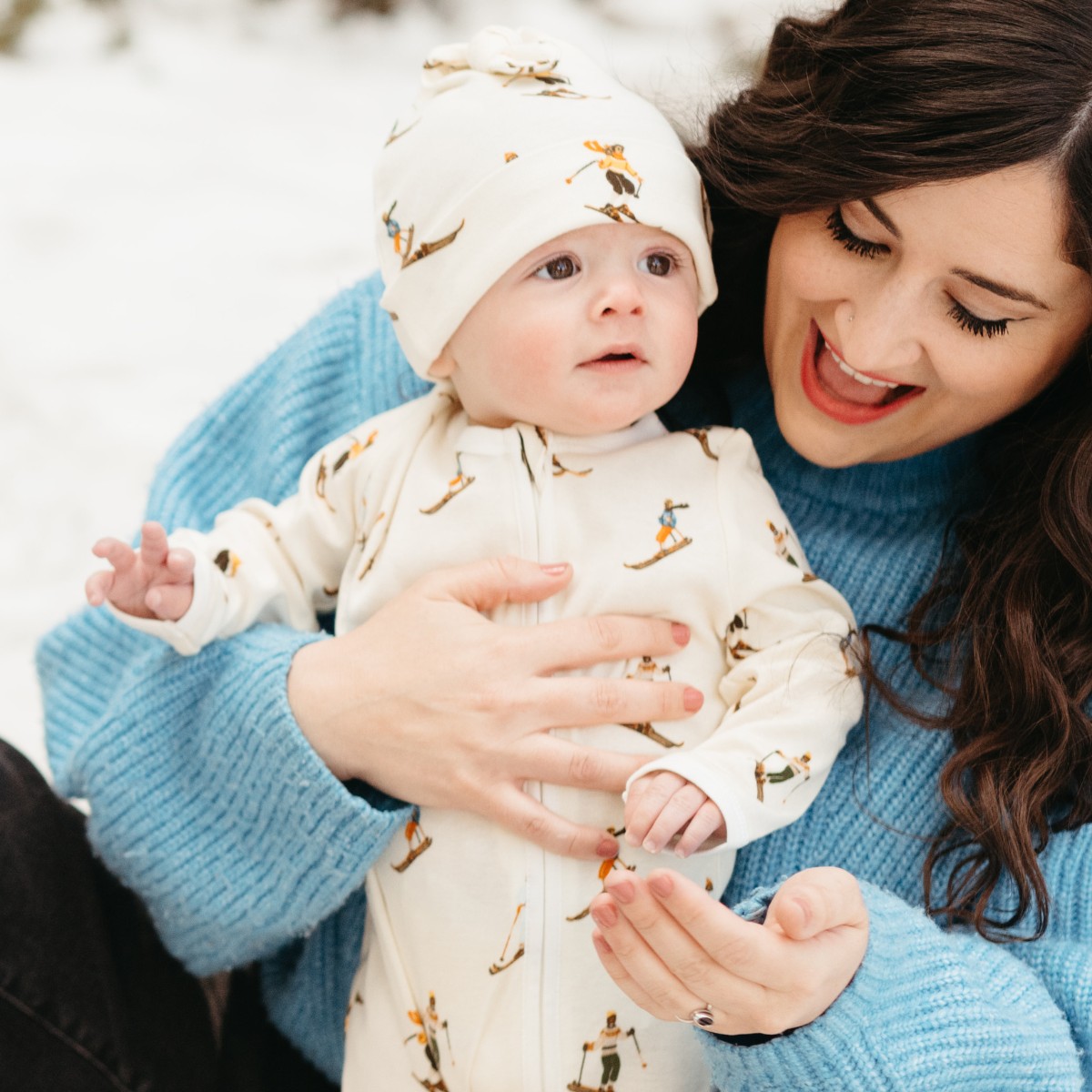 In the snow, Mom wearing a blue sweater and Baby wearing the organic cotton Vintage Natural Ski Knotted Beanie Hat and matching Zipper Footed Romper by Milkbarn Kids