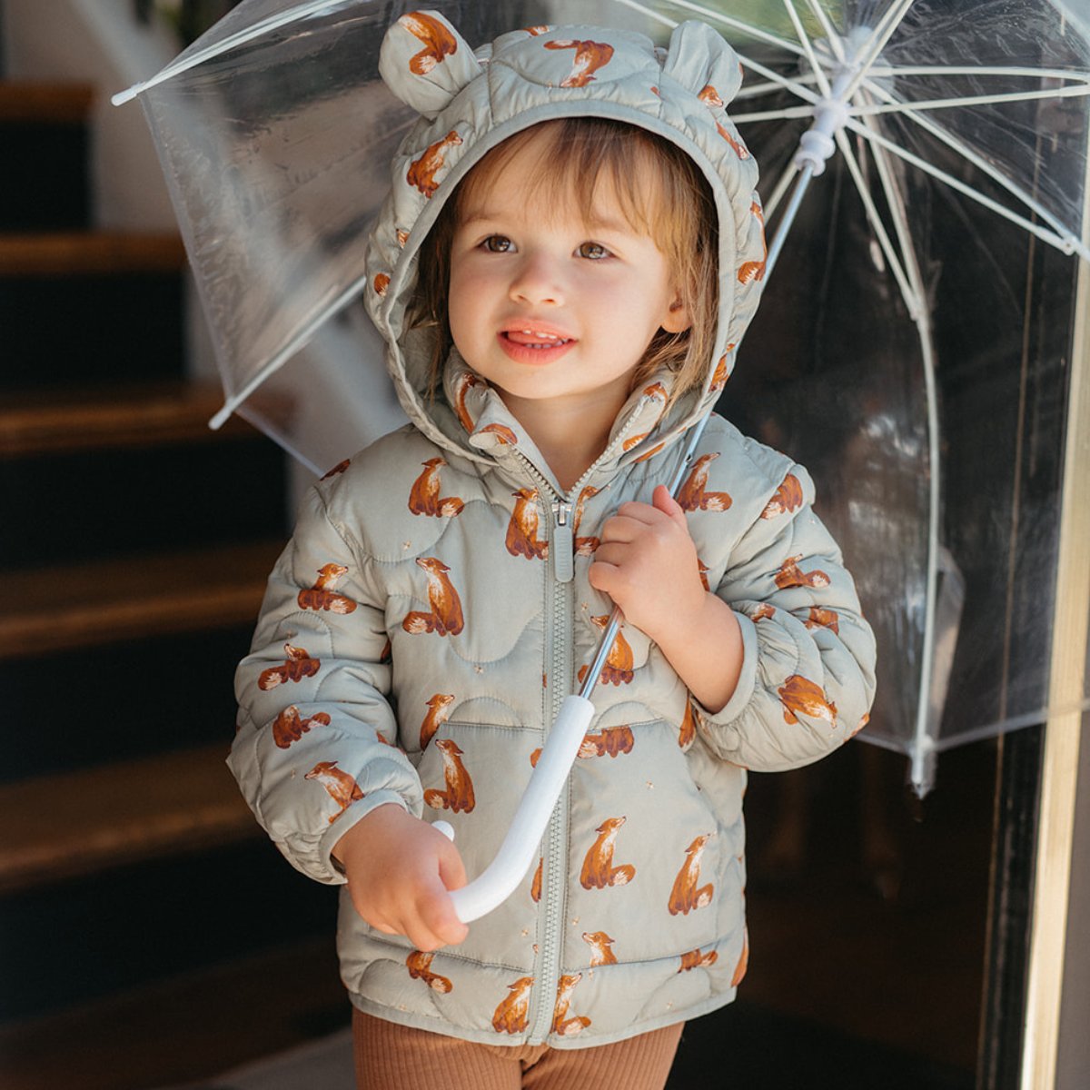 Little toddle holding a clear umbrella and standing near some stairs while wearing the Fox Lightweight Down Hooded Jacket